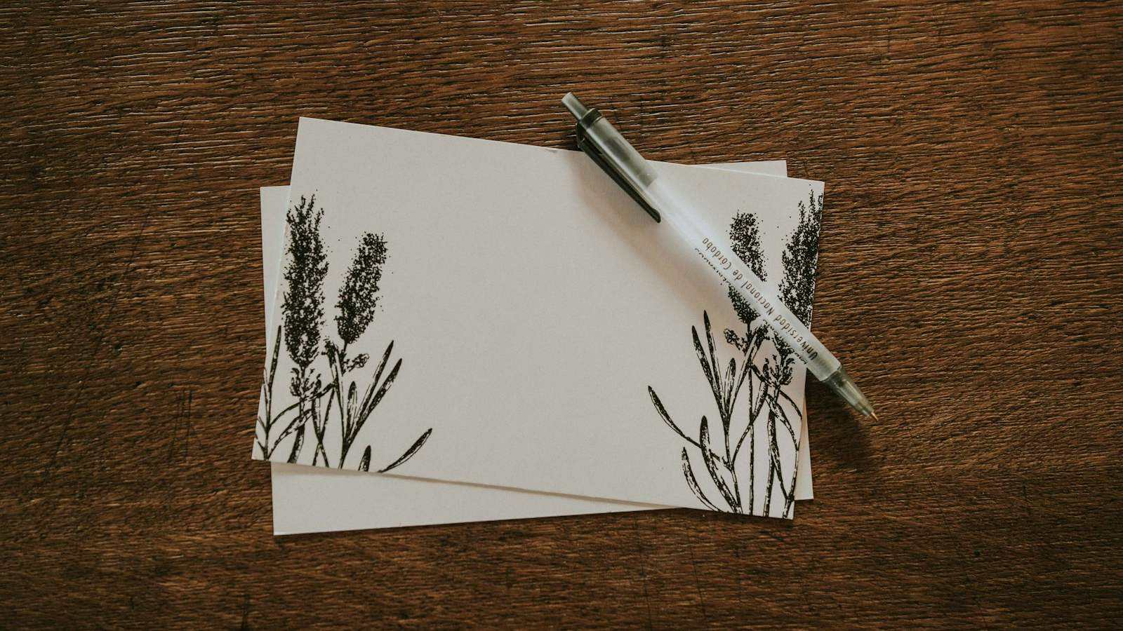 Two blank cards with a printed floral border and a pen on a dark wooden table