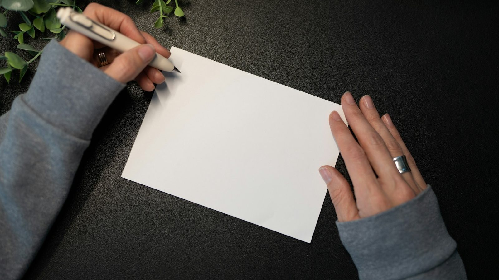 Hands holding a pen over a blank white card on a dark table, with a sprig of greenery