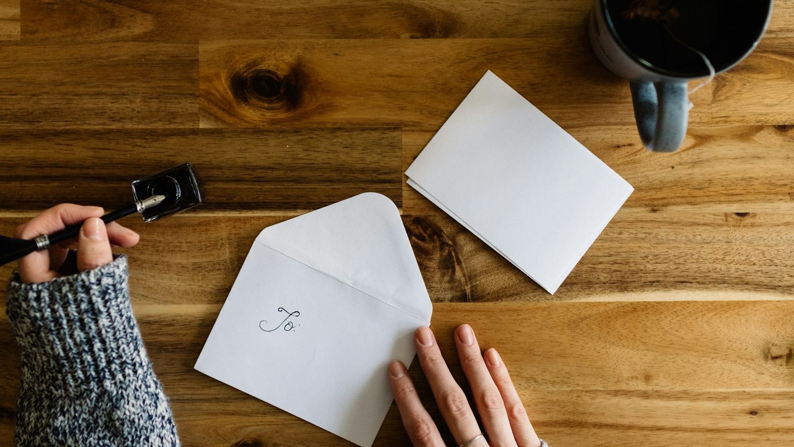 Hands writing on a white envelope at a wooden table, a card and a cup of coffee beside it